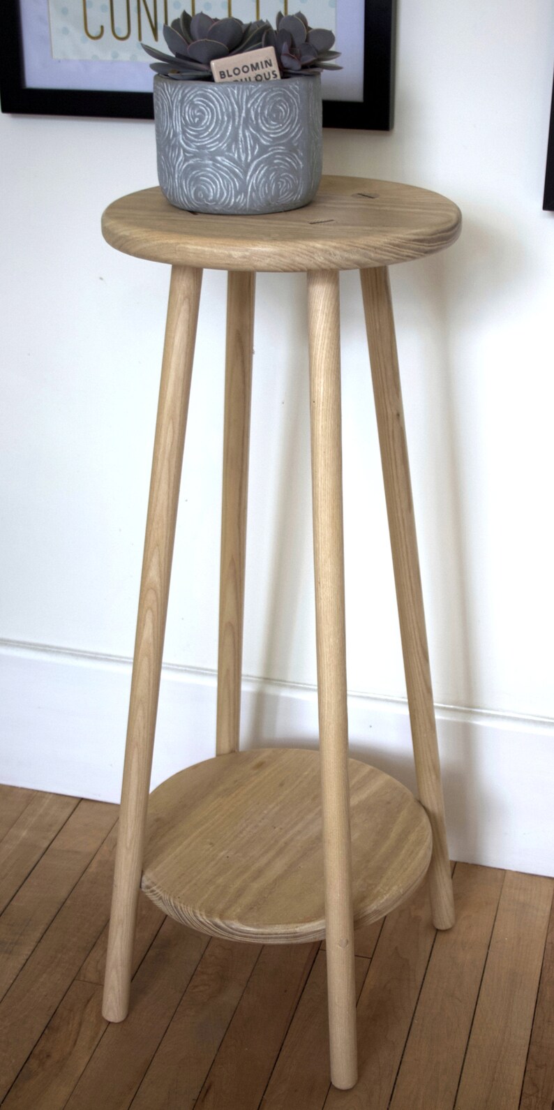 May include: A light wood plant stand with a round top and a lower shelf. The stand has four legs and is made of natural wood. A gray planter with a succulent plant is sitting on the top shelf.