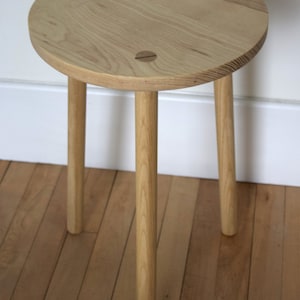 May include: A small, round wooden table with three legs. The table top is made of light wood and has a natural finish. The legs are also made of light wood and are slightly tapered.