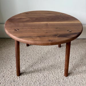 May include: A round, wooden side table with a dark walnut finish. The table has a smooth, solid surface and four slender legs. The top has decorative inlaid details. The table is approximately 18 inches in diameter and 16 inches tall.