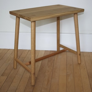 May include: A light brown wooden stool with a rectangular top and four cylindrical legs. The stool features visible joinery details on the top surface and a horizontal support bar connecting the legs near the base, constructed from natural wood.