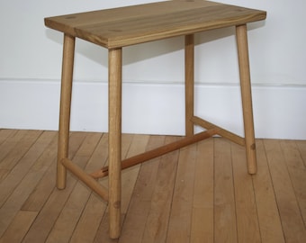 May include: A light brown wooden stool with a rectangular top and four cylindrical legs. The stool features visible joinery details on the top surface and a horizontal support bar connecting the legs near the base, constructed from natural wood.