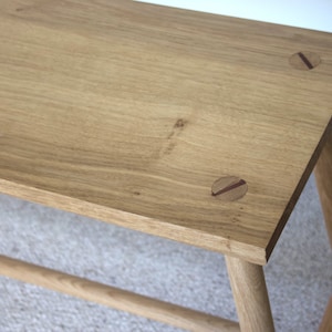 May include: A light brown wooden bench featuring a rectangular top with visible wood grain and two circular inlaid dark wood accents. The bench is supported by four cylindrical legs connected by a horizontal stretcher bar near the base.