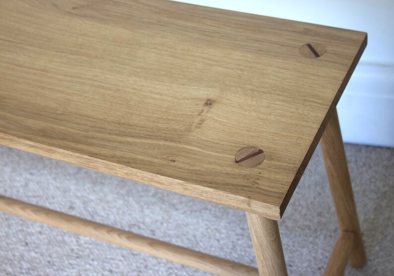 May include: A light brown wooden bench featuring a rectangular top with visible wood grain and two circular inlaid dark wood accents. The bench is supported by four cylindrical legs connected by a horizontal stretcher bar near the base.