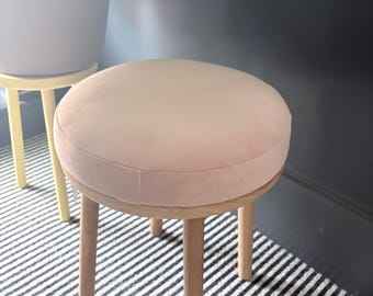 May include: A round stool featuring a soft, pale pink upholstered seat cushion supported by four light-colored wooden legs. The stool has a simple, functional design with a smooth, circular top and straight, sturdy legs made of natural wood.