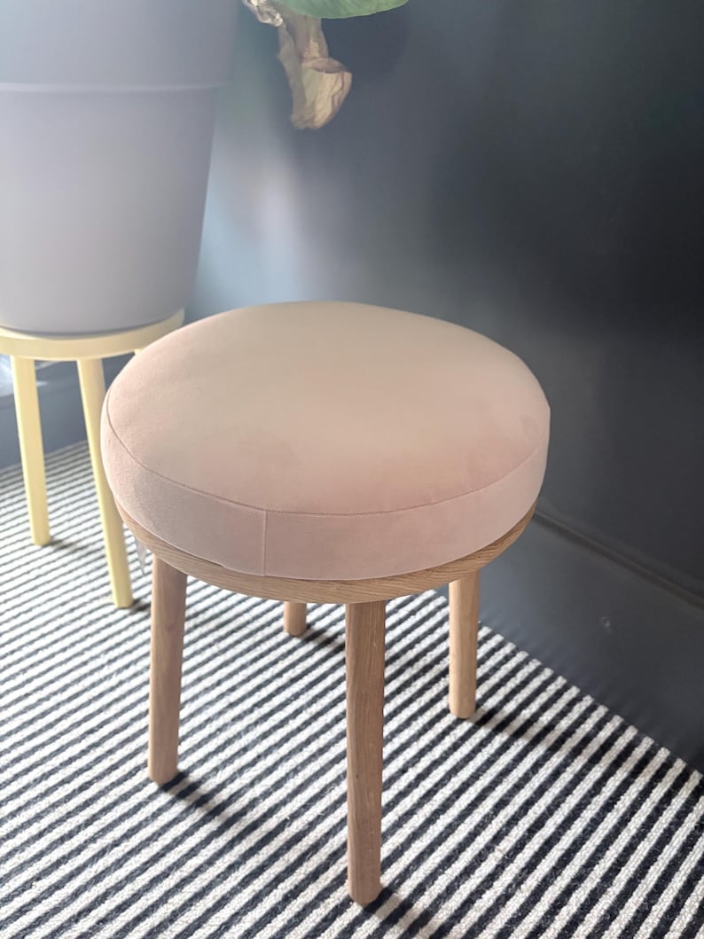 May include: A round stool featuring a soft, pale pink upholstered seat cushion supported by four light-colored wooden legs. The stool has a simple, functional design with a smooth, circular top and straight, sturdy legs made of natural wood.