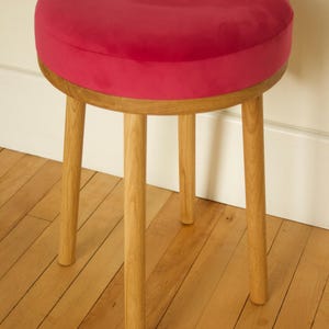 May include: A round stool featuring a vibrant magenta velvet upholstered seat cushion supported by four light-toned wooden legs. The circular seat is thick and plush, while the four cylindrical legs are positioned at an angle to provide stability.