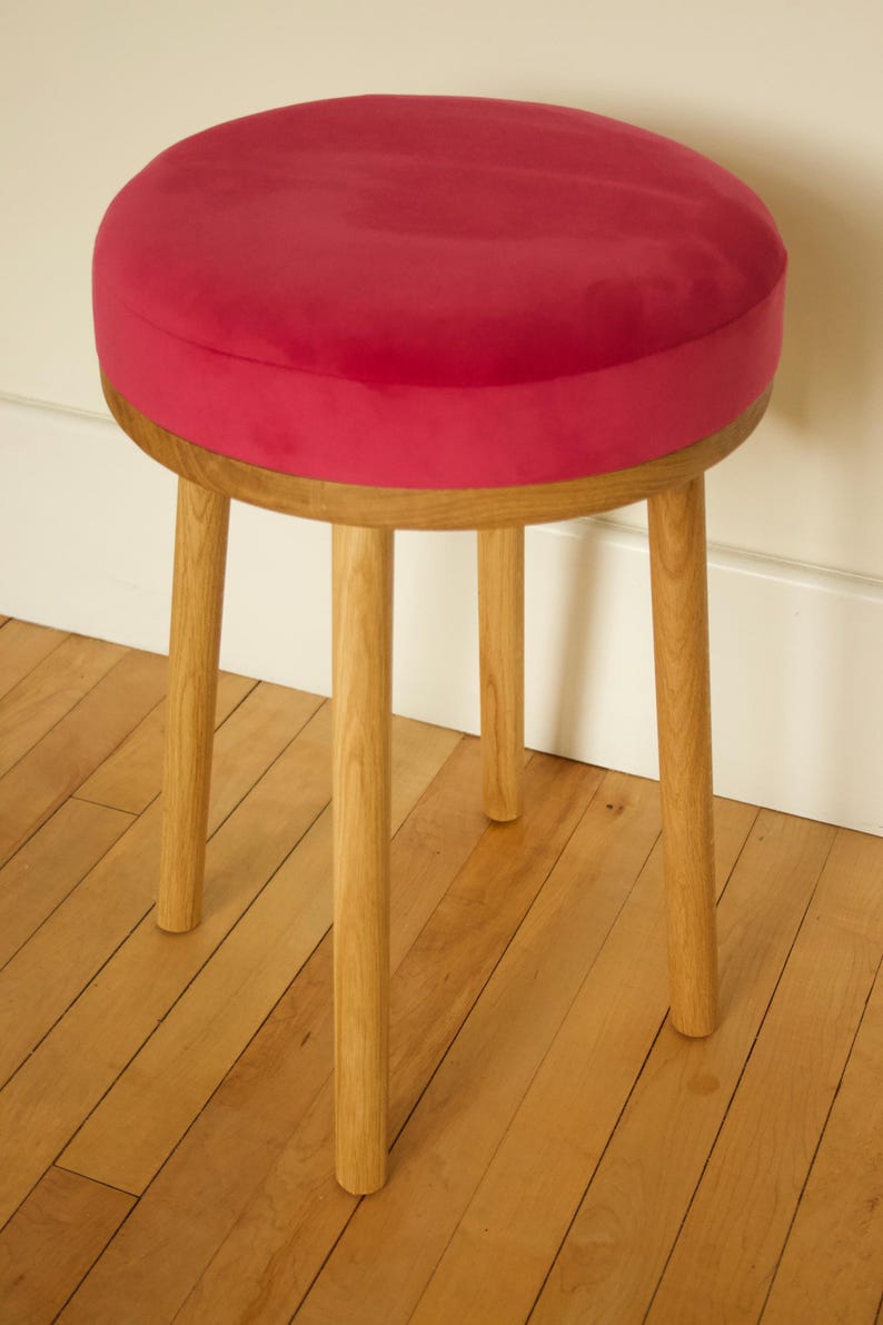 May include: A round stool featuring a vibrant magenta velvet upholstered seat cushion supported by four light-toned wooden legs. The circular seat is thick and plush, while the four cylindrical legs are positioned at an angle to provide stability.