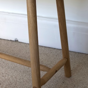 May include: A light brown wooden bench featuring a rectangular top with visible grain and four cylindrical legs. The legs are connected by horizontal support bars on the sides, and the construction includes visible circular wooden joinery details.