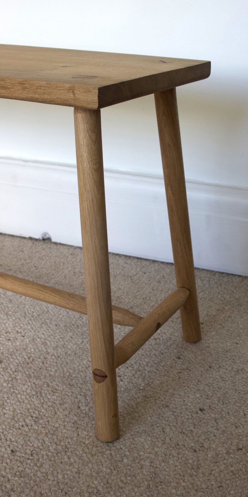 May include: A light brown wooden bench featuring a rectangular top with visible grain and four cylindrical legs. The legs are connected by horizontal support bars on the sides, and the construction includes visible circular wooden joinery details.