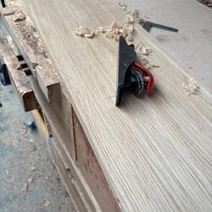 May include: A small black metal hand plane with a red adjustment lever resting on a long, light-coloured wooden board. The surface of the wood shows a natural grain pattern, and scattered wood shavings are visible around the tool.