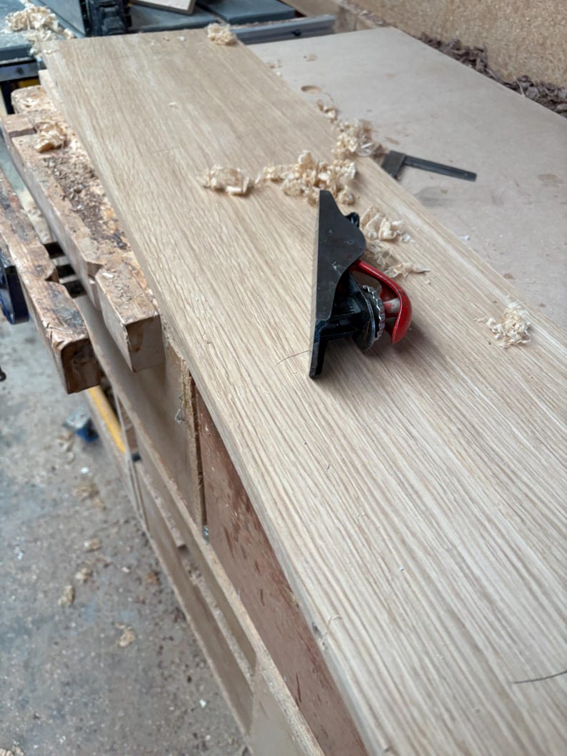 May include: A small black metal hand plane with a red adjustment lever resting on a long, light-coloured wooden board. The surface of the wood shows a natural grain pattern, and scattered wood shavings are visible around the tool.