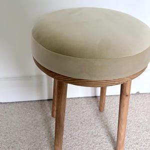 May include: A round stool featuring a beige upholstered seat with a smooth, matte texture. The seat is supported by a circular wooden frame and four cylindrical, light-toned wooden legs that extend outward from the base of the seat.