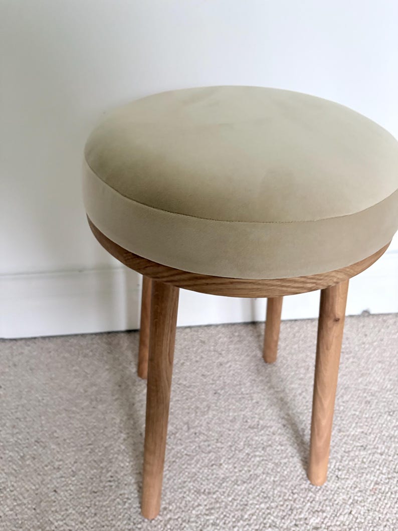 May include: A round stool featuring a beige upholstered seat with a smooth, matte texture. The seat is supported by a circular wooden frame and four cylindrical, light-toned wooden legs that extend outward from the base of the seat.
