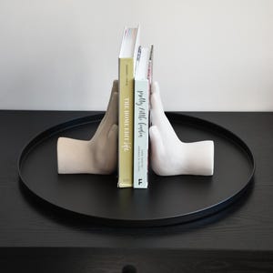 May include: A pair of hand-shaped bookends in beige and white, holding two books on a black tray. The books are titled "The Home Edit Life" and "Pretty Little Living". The tray sits on a black wooden surface.