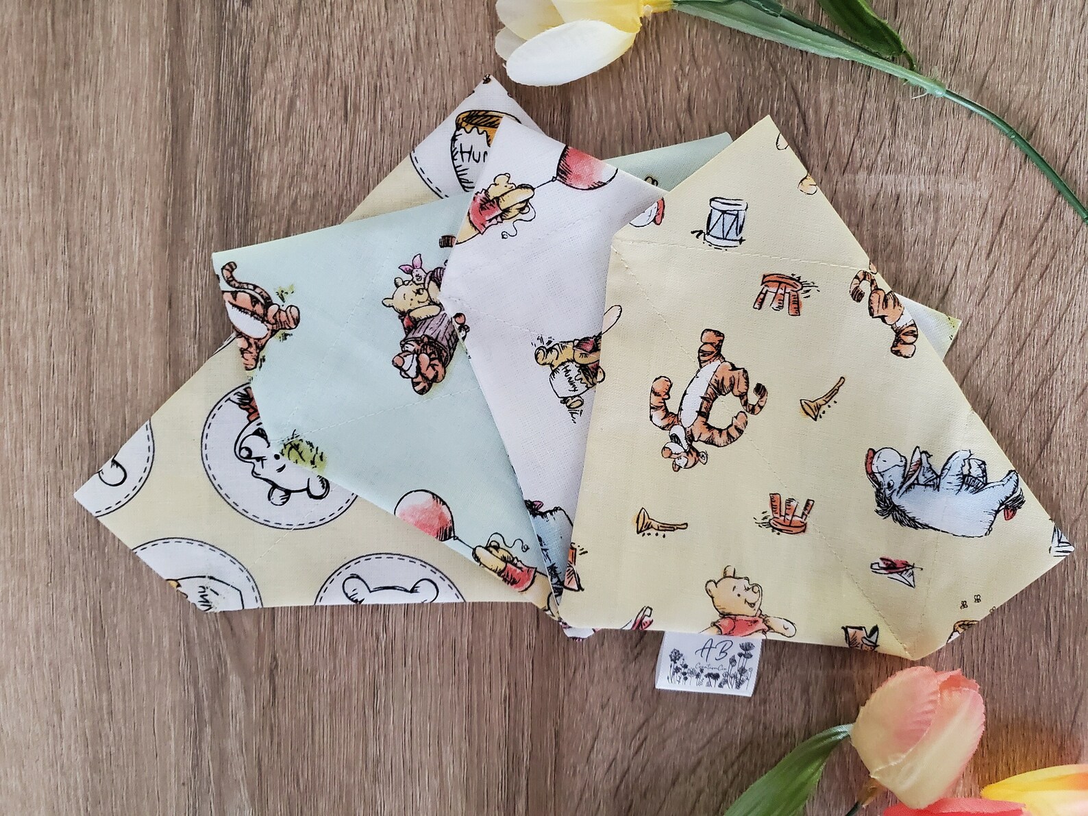 Three pastel-colored fabric squares with illustrated with "Winnie the Pooh" patterns lie on a textured wooden surface.