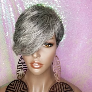 Puede incluir: Una peluca corta de color gris con raya lateral y flequillo. La peluca está peinada con un corte pixie y tiene un aspecto natural ligeramente texturizado.