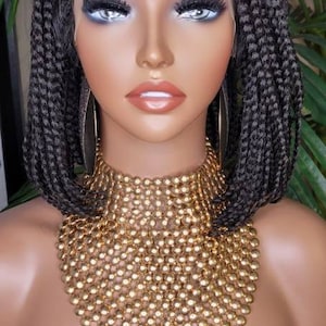 May include: A black braided wig with a lace front. The wig is styled in a bob with the braids extending past the shoulders. The wig is worn on a mannequin head with a gold beaded necklace.