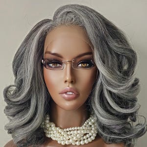 May include: A mannequin head with a gray and white wig styled in voluminous curls. The mannequin wears rimless glasses and a multi-strand pearl necklace. The skin tone is a warm brown, and the lips are a soft pink.