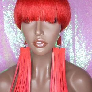May include: A red wig with a short, blunt cut and bangs. The wig is styled in a bowl cut with a straight fringe. The wig is worn on a mannequin with red fringe earrings.