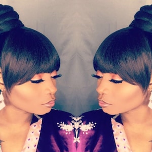 May include: A woman with dark brown hair styled in a high bun with side swept bangs. She is wearing a purple and pink floral robe and has on white earrings.