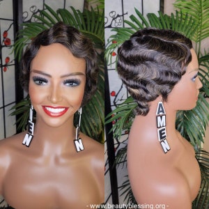 May include: A short, wavy, black and brown wig with a side part. The wig is styled in a vintage, pin-curl look. The wig is on a mannequin head with a black and white earring that says "AMEN" hanging from the right ear.