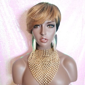 Puede incluir: Una peluca corta y entrecortada con reflejos rubios y raíces oscuras. La peluca está peinada con raya lateral y flequillo. La peluca la lleva puesta un maniquí con pendientes de aro dorados y un collar de cadena dorada.