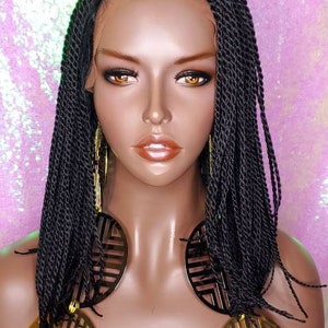 May include: A mannequin head with black braided hair and gold-colored earrings. The earrings are large, circular, and feature a geometric design. The mannequin has a tan complexion and is set against a pink, sparkly background.