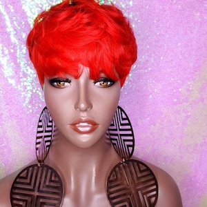 May include: A red pixie wig with a short, choppy style. The wig is styled with a side part and has a natural, wavy texture. The wig is worn by a mannequin with brown skin and is accessorized with large, circular earrings and a necklace.