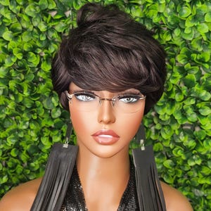 May include: A woman with short, dark brown hair is wearing a black mesh top, black leather fringe earrings, and clear-framed glasses. She is posing against a backdrop of green foliage.