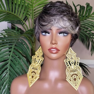 Könnte beinhalten: Eine Mannequin mit kurzen, grauen und weißen Haaren trägt große, goldene, geometrische Ohrringe. Die Ohrringe sind kunstvoll gearbeitet und haben eine dreieckige Form.