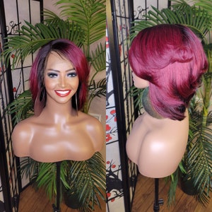 Op de afbeelding: Een kort, recht bob-pruik met een scheiding aan de zijkant. De pruik is een mix van zwart en bordeauxrood haar. De pruik is gestyled met een scheiding aan de zijkant en het haar is aan de uiteinden naar binnen gekruld. De pruik zit op een mannequinhoofd.