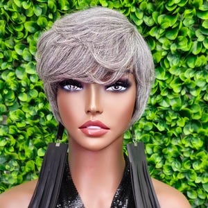 May include: A mannequin head with a short, layered, gray wig. The mannequin wears large, black leather fringe earrings and a black sequined top. The background is a wall of green foliage.