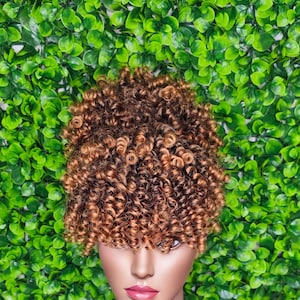May include: A brown and blonde curly hair topper with a fringe. The hair topper is on a mannequin head.