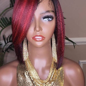 Può includere: Una testa di manichino che mostra una parrucca corta e a strati con riflessi bordeaux e neri. Il manichino è adornato con grandi orecchini a catena dorata e una collana a rete dorata. Lo sfondo include vegetazione e un muro neutro.