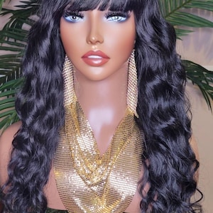 Könnte beinhalten: Eine schwarze Perücke mit lockeren Wellen und einem geraden Pony. Die Perücke ist auf einem Mannequin-Kopf mit einer goldenen Kettenkette und Ohrringen gestylt.