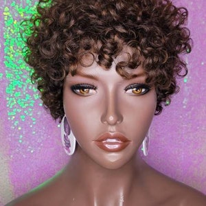 Op de afbeelding: Een bruin krullend pruik met pony op een mannequinhoofd. De mannequin heeft een bruine huid en draagt ​​oorbellen.