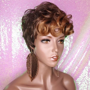 May include: A short, curly brown wig with blonde highlights. The wig is styled with a side part and has a feathered look. The wig is on a mannequin head with a pink background.