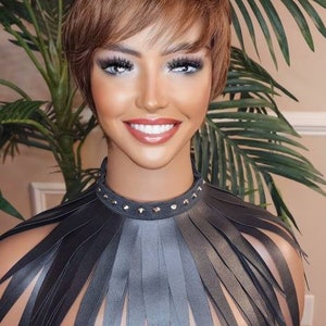 Może przedstawiać: Manekin z czarnym skórzanym kołnierzem ze srebrnymi ćwiekami i długimi, postrzępionymi krawędziami. Manekin ma krótkie brązowe włosy i jest ustawiony na tle zielonych liści palmowych.