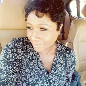 May include: A woman with short, curly black hair is sitting in a car. She is wearing a black and white floral dress.