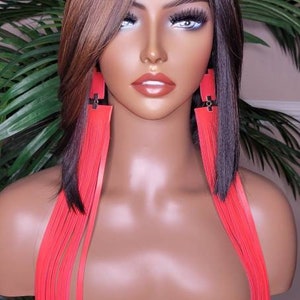 Może przedstawiać: Mannequin z długimi, brązowymi i blond włosami w długich, czerwonych kolczykach z frędzlami. Mannequin ma na sobie czarną bluzkę i stoi przed zieloną rośliną.