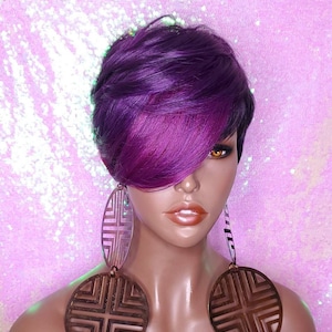 Puede incluir: Una peluca corta de estilo pixie, de color morado y magenta, con raya lateral. La peluca está peinada con un flequillo lateral. La peluca está en una cabeza de maniquí con grandes pendientes circulares geométricos.