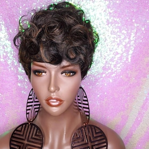 Puede incluir: Una peluca corta y rizada de color marrón y rubio con raya lateral. La peluca está peinada con un corte pixie y la lleva un maniquí con grandes pendientes circulares.