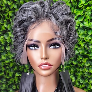 May include: A mannequin head with a short, curly, gray wig. The wig has a lace front. The mannequin is wearing large, black, fringed earrings and a black sequined top. The background is a wall of green leaves.