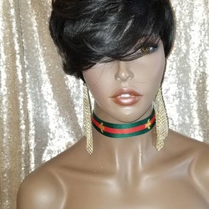 Puede incluir: Una peluca corta negra con raya lateral y flequillo. La peluca está peinada con un corte pixie. La peluca está en una cabeza de maniquí con un collar de gargantilla de rayas verdes y rojas con detalles dorados.