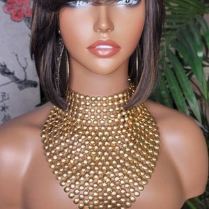 Puede incluir: Una cabeza de maniquí con una peluca bob de color marrón y rubio con flequillo de lado. El maniquí lleva un collar de cuentas doradas.