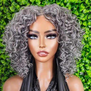 May include: A mannequin head with a short, curly, gray wig. The wig has a natural-looking hairline and is styled with voluminous curls. The mannequin has full makeup, including dark eyelashes and a neutral lip color. The background is a wall of green leaves.