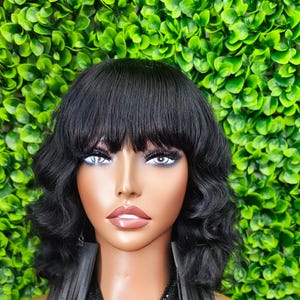 May include: A black wig with a blunt fringe and wavy ends. The wig is styled on a mannequin with a fair complexion, against a backdrop of green foliage. The wig's texture appears smooth and the colour is a deep, solid black.