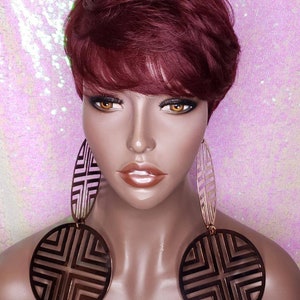 Puede incluir: Un maniquí con el pelo corto de color rojo burdeos que lleva grandes pendientes geométricos de color dorado. Los pendientes están hechos de varias formas circulares y ovaladas con un diseño recortado.