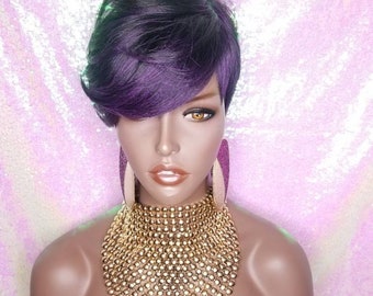 May include: A short, purple and black wig with side-swept bangs. The wig is styled in a pixie cut and is worn on a mannequin head. The mannequin is wearing a gold necklace.