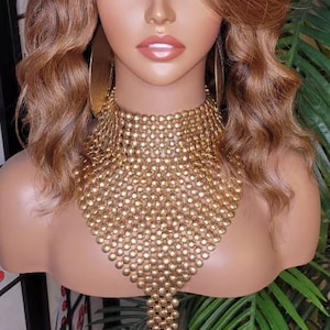 Könnte beinhalten: Eine Mannequin mit langen, welligen, hellbraunen Haaren mit blonden Strähnen, die eine goldene Kettenkette trägt. Die Mannequin trägt eine goldene Halskette und große goldene Ohrringe.